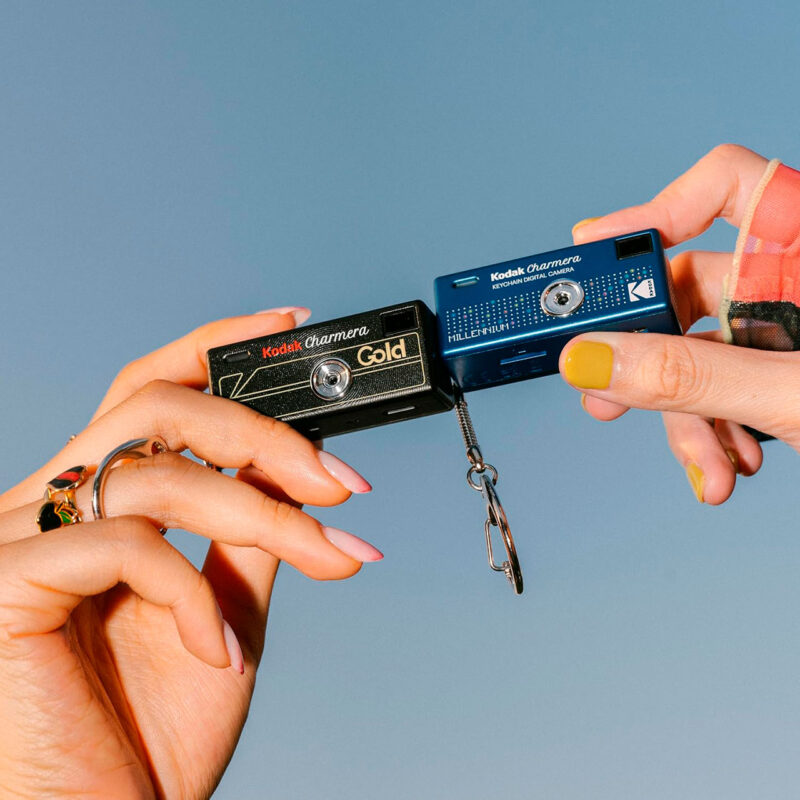 kod200091-3-8cb Two Kodak Charmera cameras, one black Gold and one Millennium blue, held by hands, bright blue background.