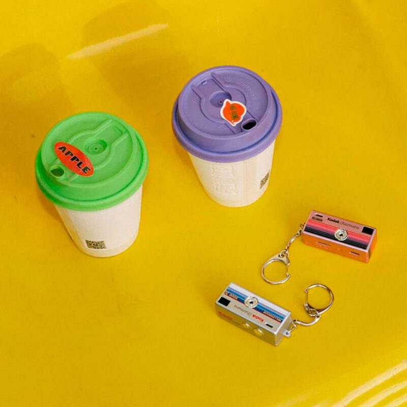kod200091-4-839 Drink glasses with green and purple lid next to Kodak Champion camera keychains on yellow background.