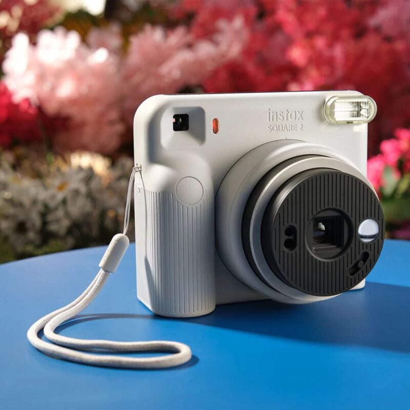 fuj170532-6-04a Cámara instantánea Fujifilm Instax Square SQ2 de color blanca, con lente y flash, sobre fondo azul.